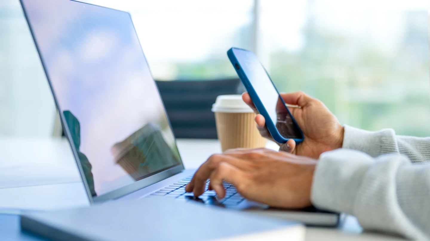 Close up of a Businessman working on a laptop computer and holding and looking at a mobile phone in the office. There are notepads on the desk. He is casually dressed