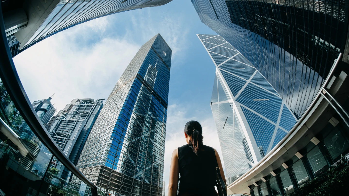 Rear view of professional young businesswoman standing against contemporary financial skyscrapers in downtown financial district and looking up into sky with positive emotion