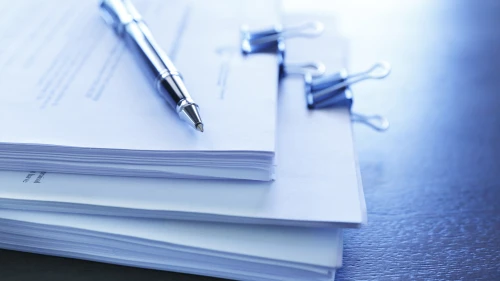 A ballpoint pen rests on top of a stack of documents ready for signing. The image is photographed using a very shallow depth of field with the focus being on the tip of the pen.