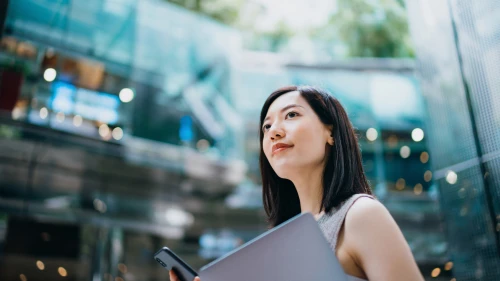 Successful and modern young Asian businesswoman carrying smartphone and laptop, commuting to work in central business district against contemporary corporate buildings in the city. Female leadership. Business on the go