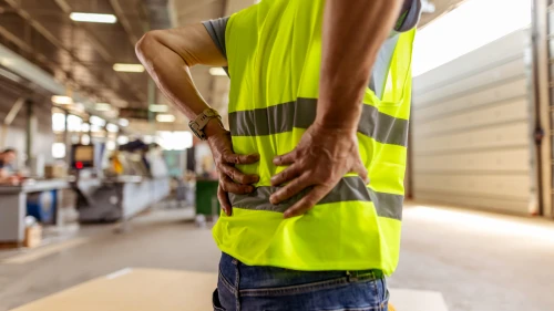 A view of a worker wearing a safety vest clutching their lower back in a factory environment. The image highlights workplace safety, physical strain, and occupational health risks in industrial settings.