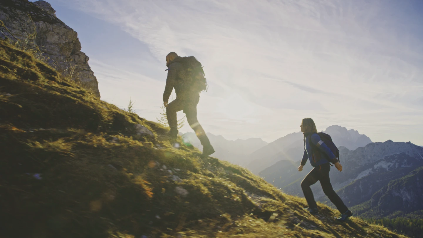 A mid adult man and woman hike along a scenic mountain trail during sunset, dressed in outdoor attire, with backpacks, embodying an active lifestyle in nature.